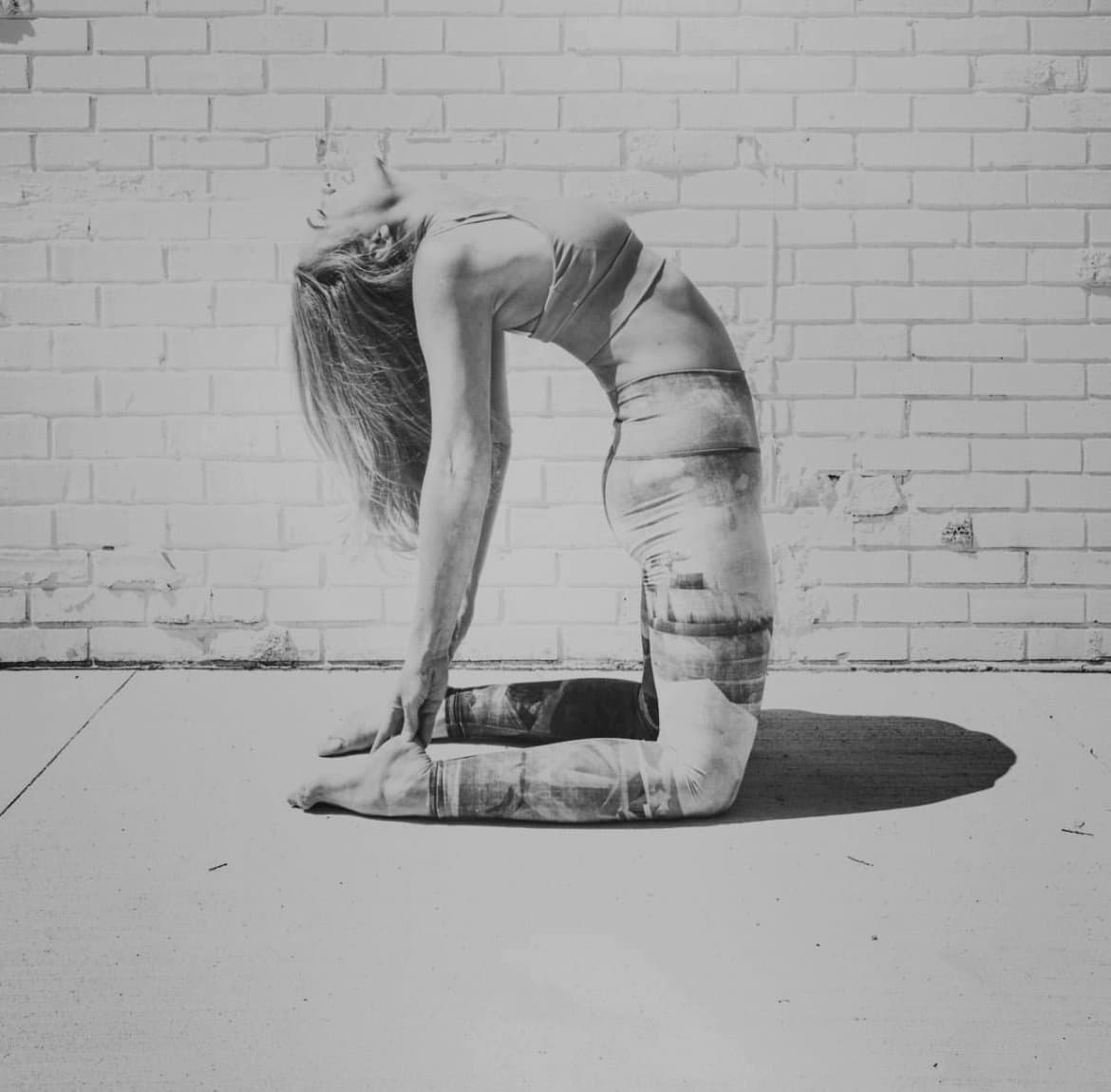 A student in a deep kneeling backbend against a white brick wall